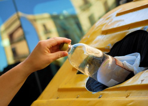 A person's hand holding a plastic bottle filled with clear liquid, positioned over a yellow waste bin with a circular opening, which contains scattered papers and other waste materials. The bin is situated outdoors on a paved surface, likely on a driveway or street area, with blurred buildings and greenery visible in the background. The scene is well-lit, suggesting daytime, and emphasizes waste disposal in a typical domestic or urban setting. This image relates to rubbish collection and waste management services that Ilford House Clearance provides for residential and commercial clients in Ilford and nearby regions.