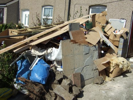A large pile of mixed waste materials situated on a pavement area outside a residential building in an urban setting. The heap includes various types of rubbish such as broken concrete blocks, wooden planks, cardboard boxes, and plastic bags. Some cardboard boxes are flattened or partially collapsed, while others are still intact. There are also pieces of plasterboard and insulation material visibly incorporated within the debris. The waste appears to be haphazardly piled, with items overlapping and scattered across the sidewalk, adjacent to the exterior wall of a building with two windows. The surroundings suggest a typical street scene in a suburb, with neighboring houses and some garden shrubbery in the background. The lighting indicates a clear day, and the overall scene reflects the kind of clutter that might require professional rubbish removal services in Ilford or nearby areas.