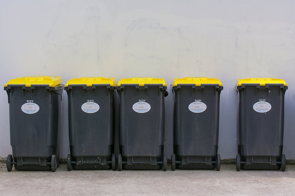 A blue plastic wheelie bin with a closed lid, positioned outdoors on a grassy area, features a yellow and black sticker that reads 'FIGHT BACK!' The bin is placed next to a red bin with a black lid, b