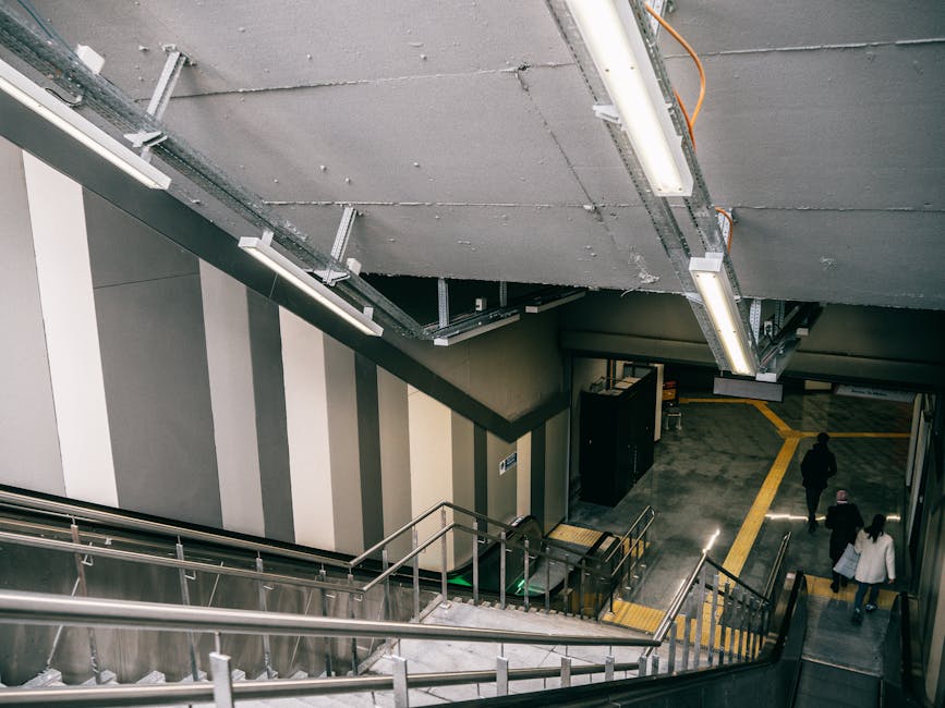 The image depicts an underground station interior from an elevated perspective, showing a staircase with metallic railings leading down to a lower level. The staircase is situated against a wall paint