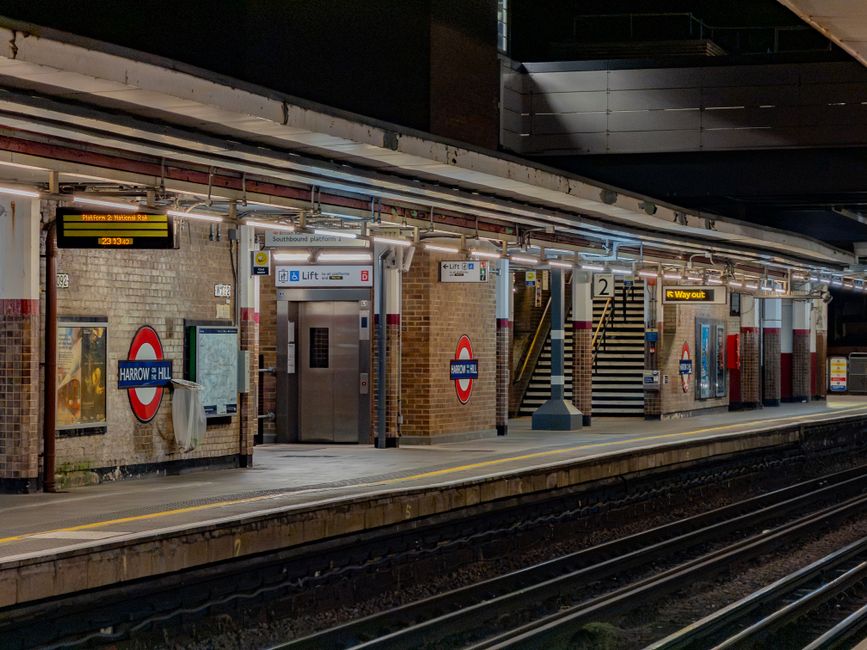 The image depicts an underground station interior from an elevated perspective, showing a staircase with metallic railings leading down to a lower level. The staircase is situated against a wall paint